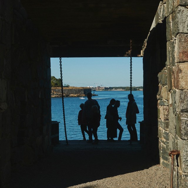 A group of four adults and 2 children stand silhouetted by the shadow of a large portcullis (Kuninkaanportti). Our point of view is from inside the fort, the walls of the fort are so thick it almost looks like a tunnel. Chains hang down from the ceiling of the entrance way and are connected to the drawbridge on which the silhouetted family stand. Behind them we can see a brightly lit open expanse of water. A small boat passes from right to left, and on the left side we can see the edge of an island, with reinforced stone walls. In the far distance we can see what appears to be the mainland with tall buldings and even taller construction cranes.