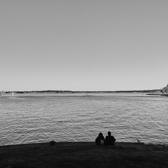 WIde-angle landscape black and white photo showing a silhouetted couple sitting beside each other on the edge of large body of water. They appear to be gazing out at the sea in front of them. Beside them a seagull sits, also appearing to observe the water. We can clearly see one small motorboat in the distance moving directly away from us. Some other small boats can be seen, along with a small glimpse of shoreline at either edge of the photo.